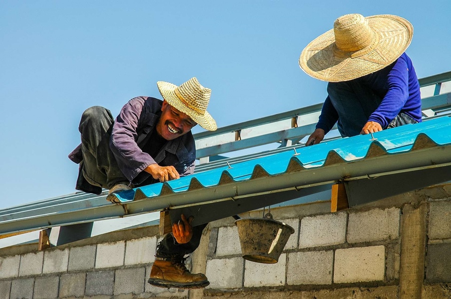 Two roofers working on a roofing project that uses blue roofing sheets who are wearing hats, boots, and long clothes to get protection from sunlight.