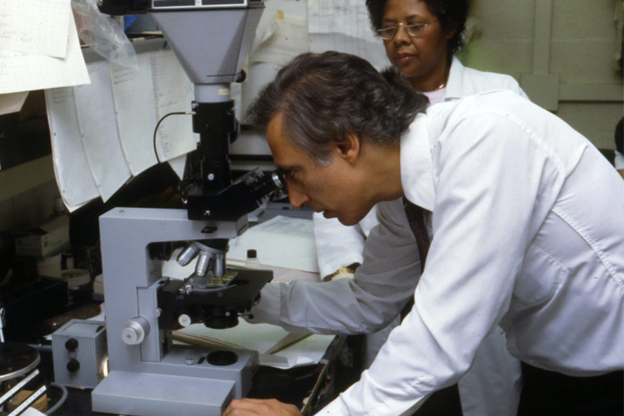 A person inspecting a specimen with another person next to him using a compound microscope, which is one of the different types of microscopes